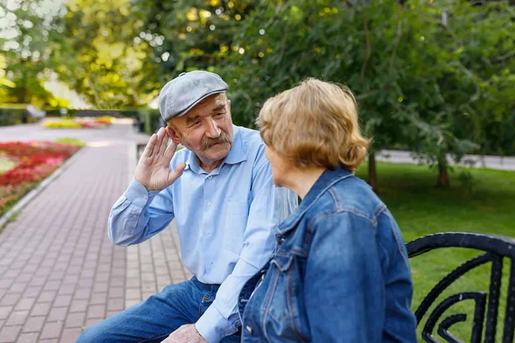 An senior man and his friend sitting in a park talking, as the man puts his hand up to his ear to hear better